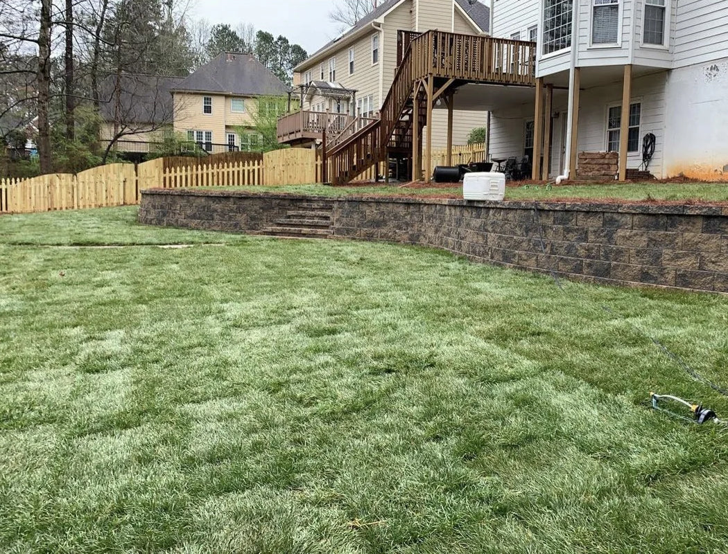 After completion: expansive level sod lawn bordered by a sweeping multi-tier natural stone retaining wall with integrated steps, creating a clean, usable backyard space under an elevated deck with professional grading, drainage, and hardscaping transformation in Marietta, GA by Precision Grading & Hardscapes