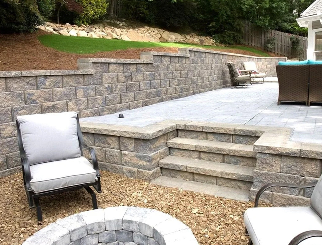 Elegant terraced retaining wall featuring textured stone blocks, built-in staircase, and expansive gray paver patio with central fire pit and lounge furniture in a sloped backyard in Marietta, Georgia