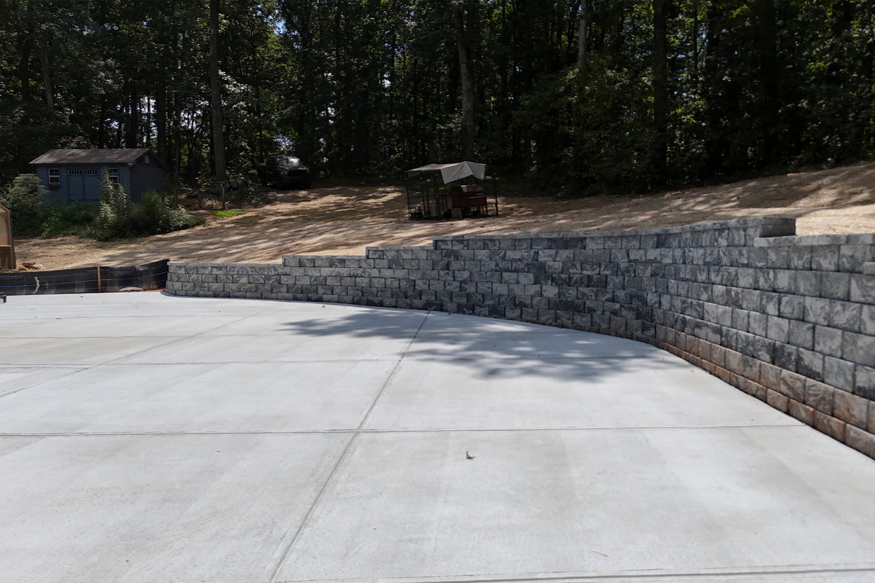 Retaining wall made of stone blocks with a concrete driveway in front, surrounded by trees.