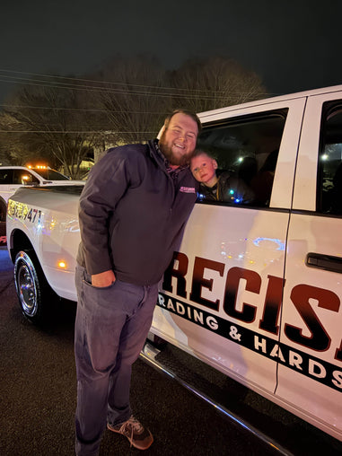 Man and child standing next to a white truck with 'PRECISION TRADING & HARDSCAPES' branding at night.
