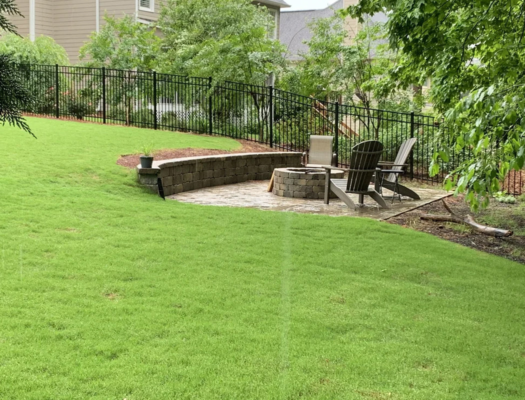 Beautifully completed backyard oasis with rich green sod, central stone fire feature, curved retaining wall seating, and tall black fence framing a peaceful wooded setting in Marietta, Georgia