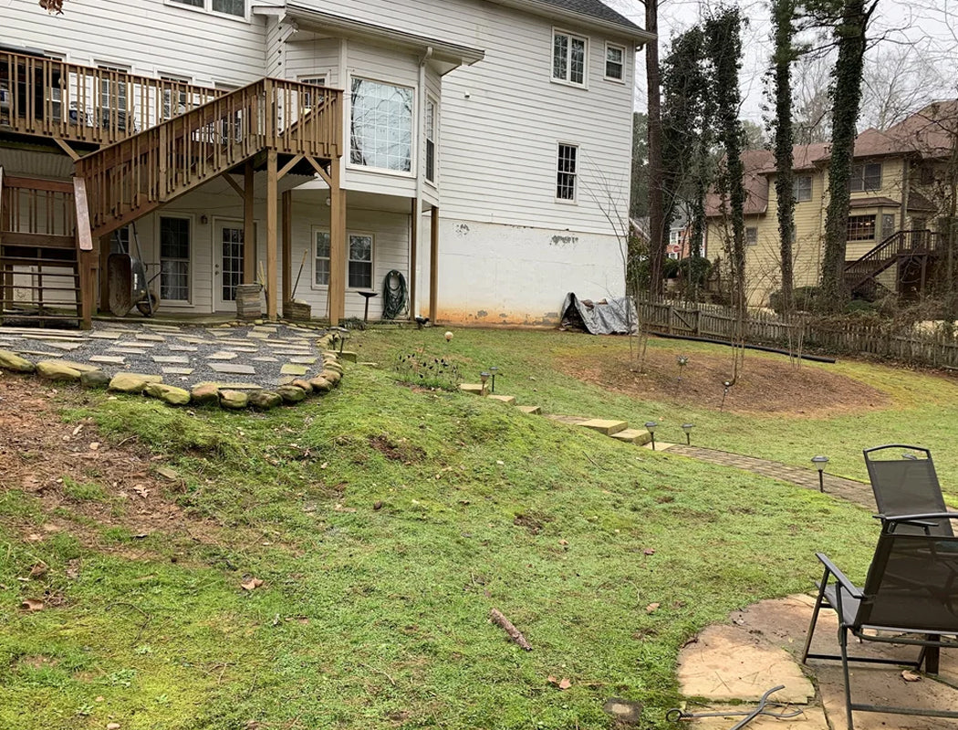 Before transformation: uneven, mossy stepping stones scattered across a sloped, patchy backyard with exposed dirt, overgrown grass, and makeshift drainage issues beneath an elevated deck with a challenging slope in need of professional grading, retaining walls, and hardscaping in Marietta, GA