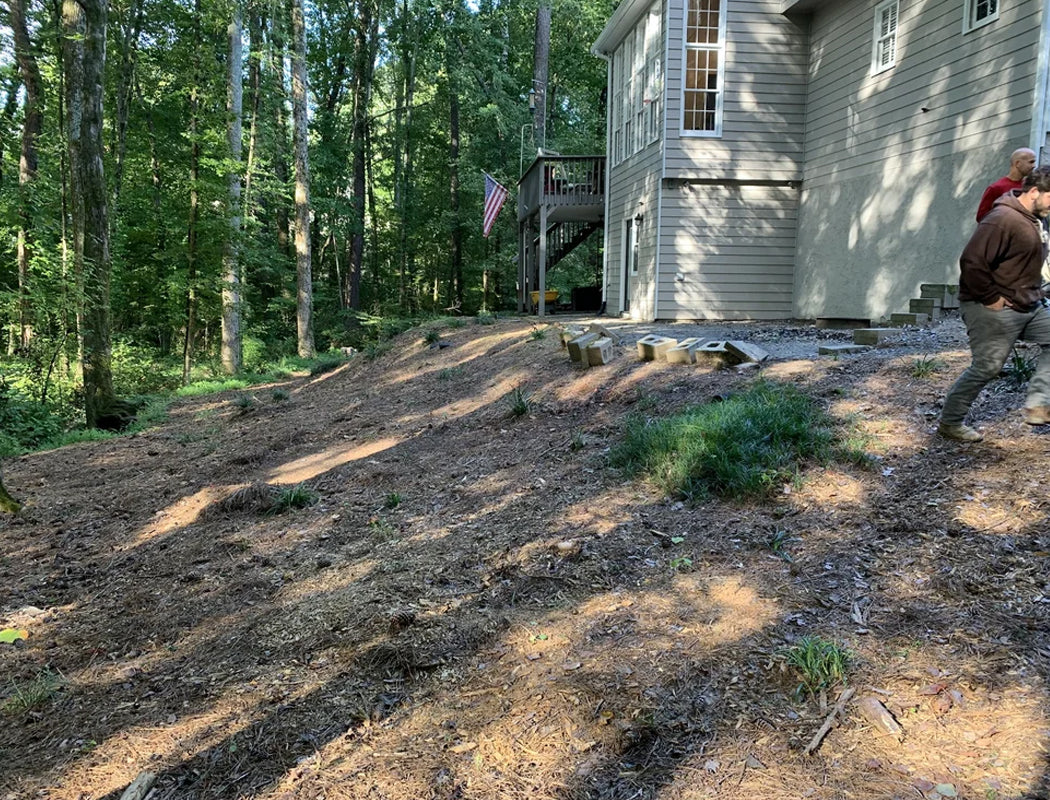 Before view: steep, uneven wooded hillside with patchy grass, scattered concrete blocks, exposed dirt, and erosion-prone slope adjacent to house foundation and deck stairs in Marietta, Ga