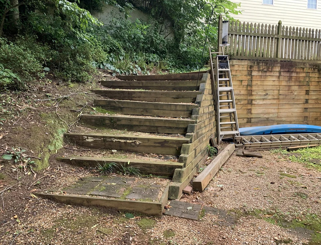 Before view: steep, uneven wooden hillside steps with deteriorating treads and makeshift side support, leading from upper yard to lower area beside existing fence