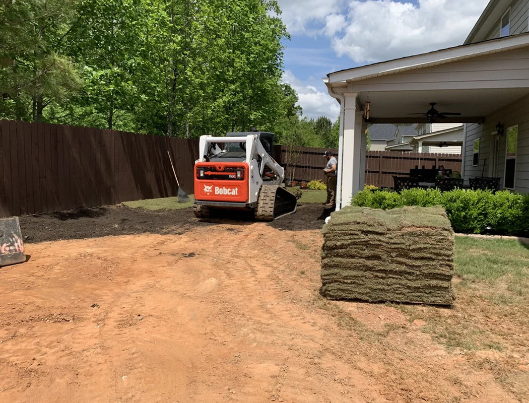 Bobcat tractor in a backyard with a pile of sod and a house in the background.