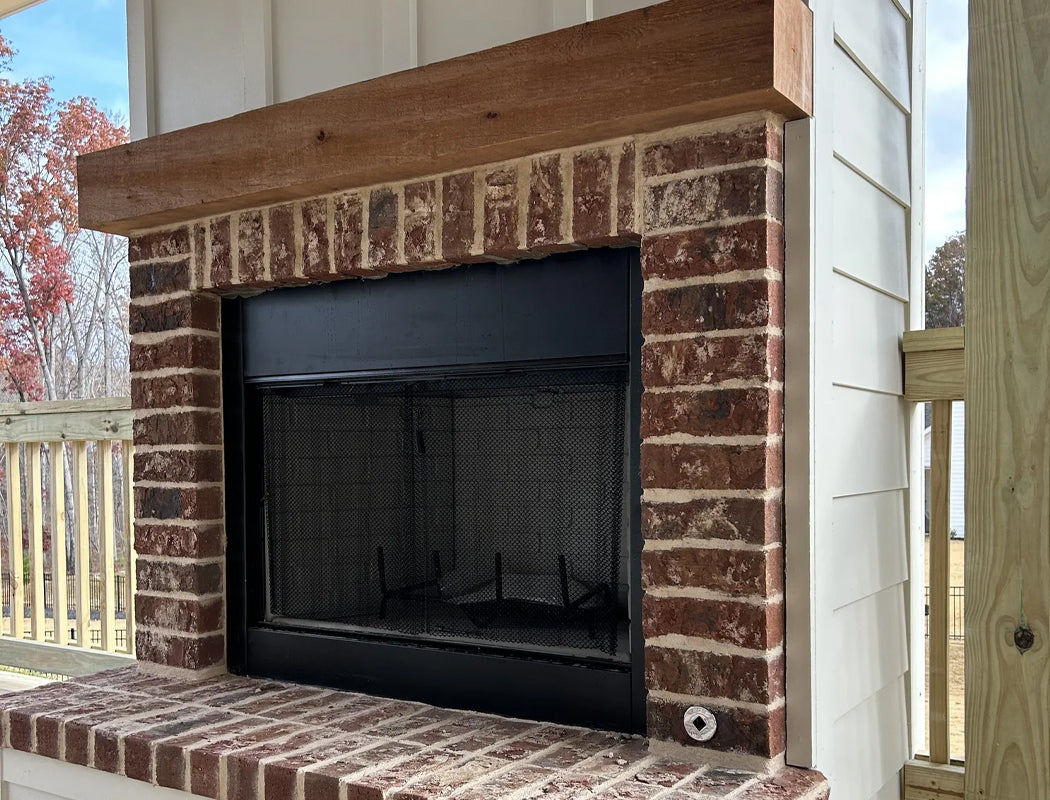 Rustic outdoor brick fireplace with wide reclaimed-wood mantel, black metal firebox insert, and clean brick surround
