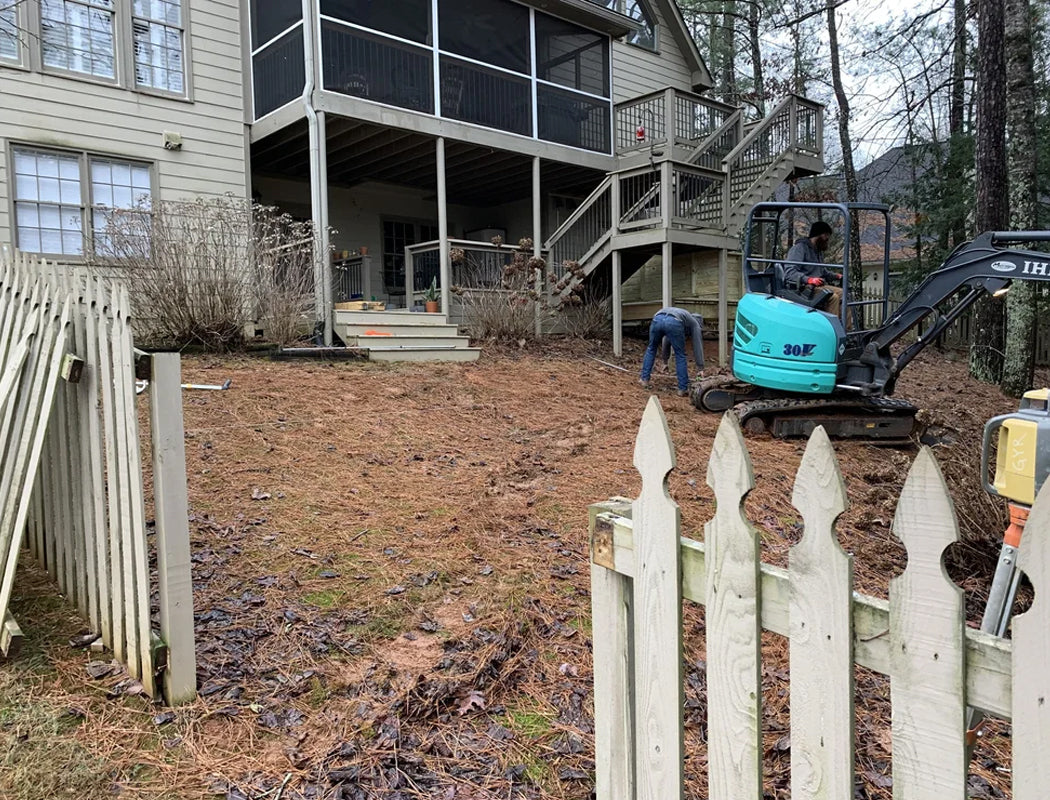 Excavator in front of a house with a white picket fence and leaf-covered ground.
