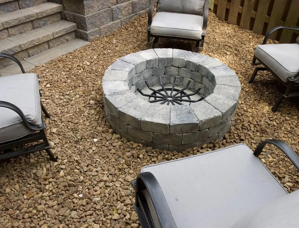 Nestled in a serene backyard corner, a handcrafted circular stone fire pit with sturdy gray block surround and central metal grate sits amid a bed of smooth pea gravel, encircled by comfortable cushioned armchairs ready for evening fireside relaxation