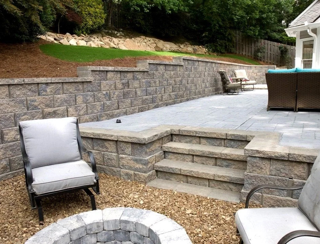 Outdoor patio with stone retaining wall, steps, and chairs in a backyard setting.