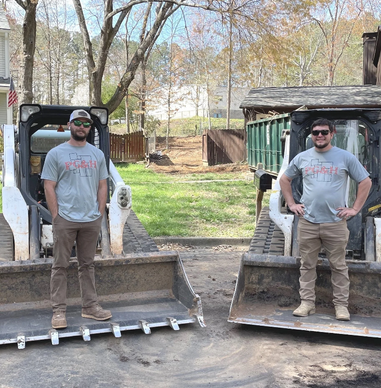 Two men standing on the buckets of two excavators in an outdoor setting.