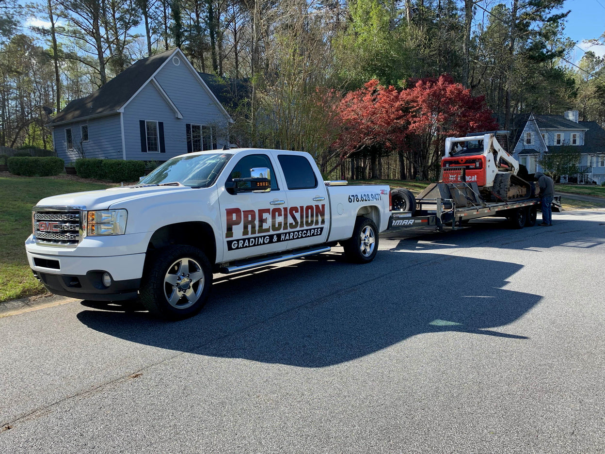 White truck with 'Precision Grading & Hardscapes' on a residential street with a skid steer in tow