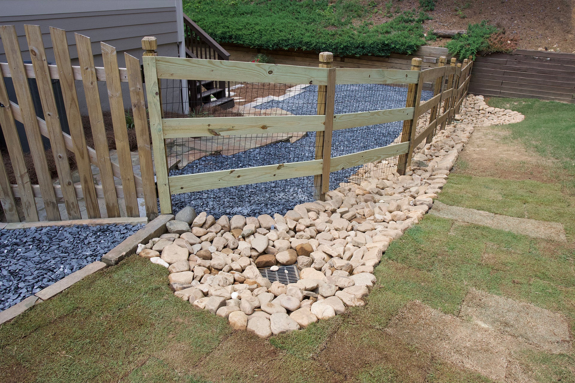 Curved wooden fence with black wire mesh infill and timber posts, seamlessly integrated with a wide gravel-and-stone drainage swale featuring a central catch basin grate to manage runoff and protect the lawn.